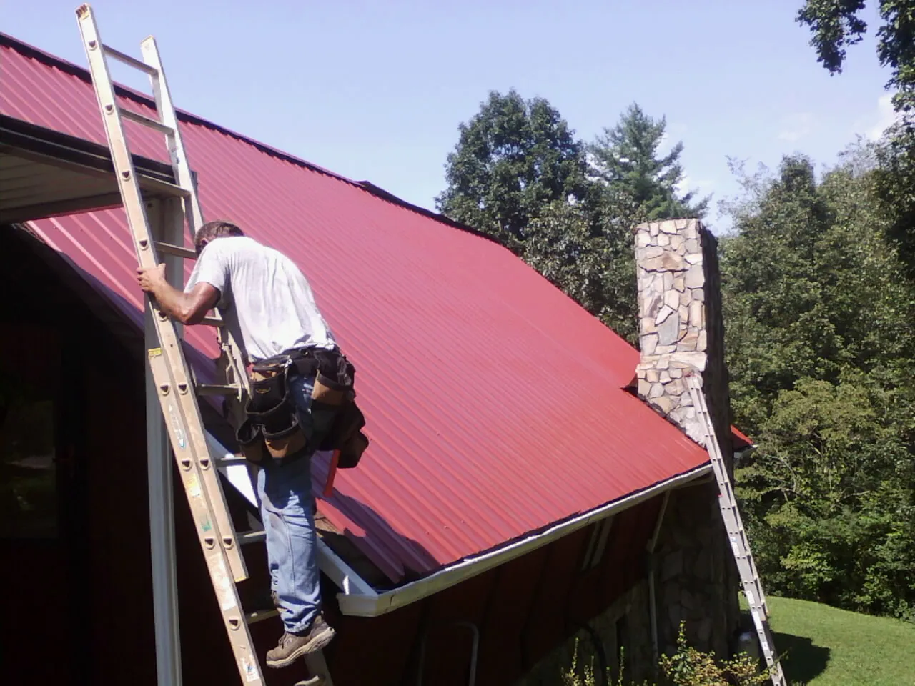 Roofer on a ladder beside a red metal roof and stone chimney — Soffit & Fascia Repair by Roofing Brothers in Mineral City